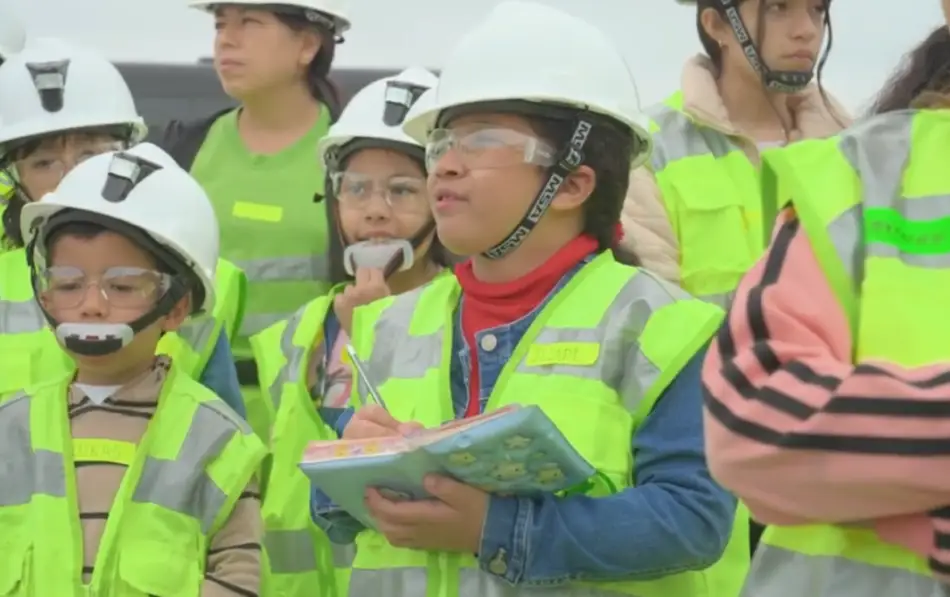 Grupo de niños o jóvenes con cascos de seguridad blancos y chalecos verde neón, prestando atención.