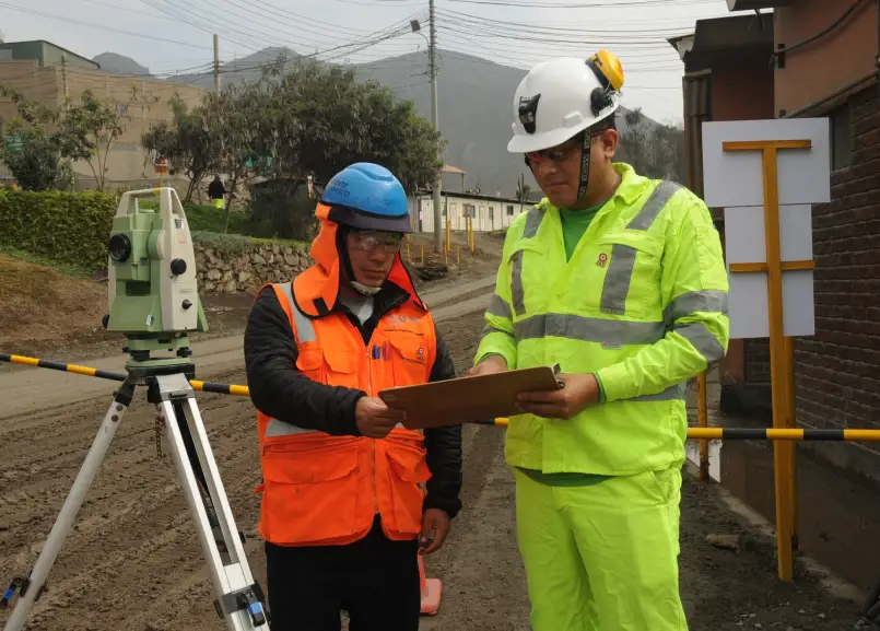 Dos colaboradores en la obra, uno con chaleco naranja y el otro con chaleco verde neón, revisando documentos junto a un equipo topográfico.